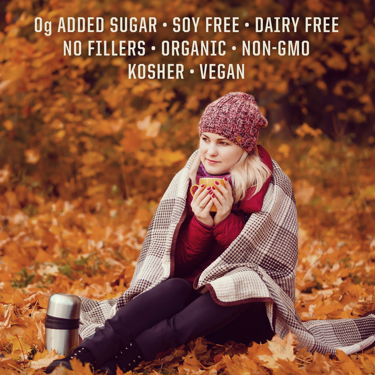 Woman wrapped in plaid blanket sitting on autumn leaves holding a warm cup beside a thermos flask
