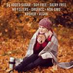 Woman wrapped in plaid blanket sitting on autumn leaves holding a warm cup beside a thermos flask