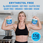 Woman flexing arms holding two containers of plant based protein powder in kitchen background