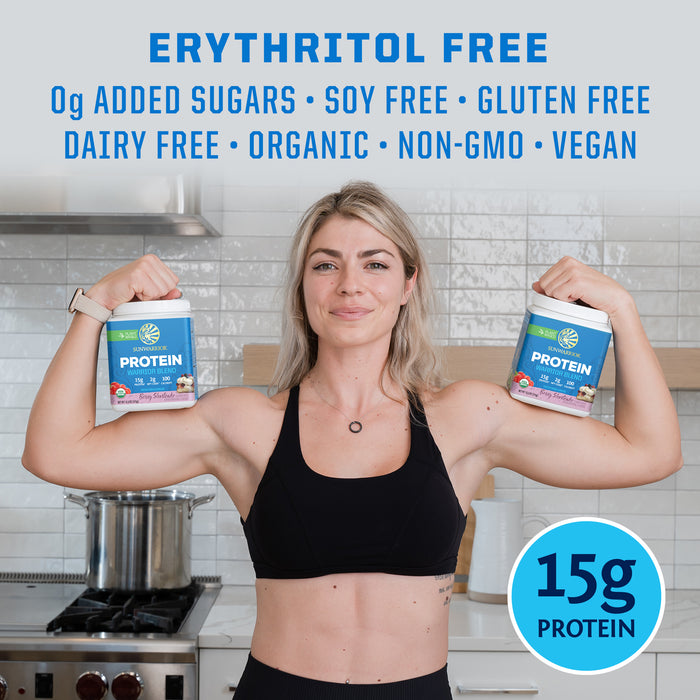 Woman flexing arms holding two containers of plant based protein powder in kitchen background