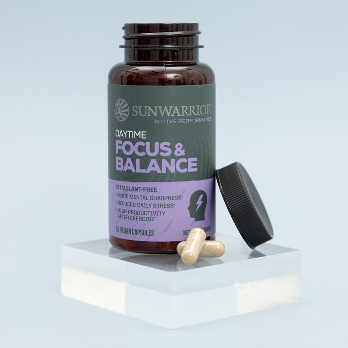 Brown bottle of Sunwarrior daytime focus and balance capsules with black lid removed, placed on a clear acrylic block with two beige capsules beside it against a light background.