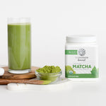 Glass of green matcha drink next to container of organic powdered matcha and a spoon with powder