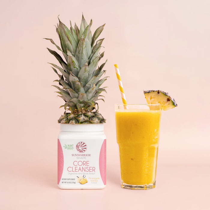 Jar of pink-labeled cleanser with pineapple top beside yellow smoothie in glass with striped straw and pineapple wedge
