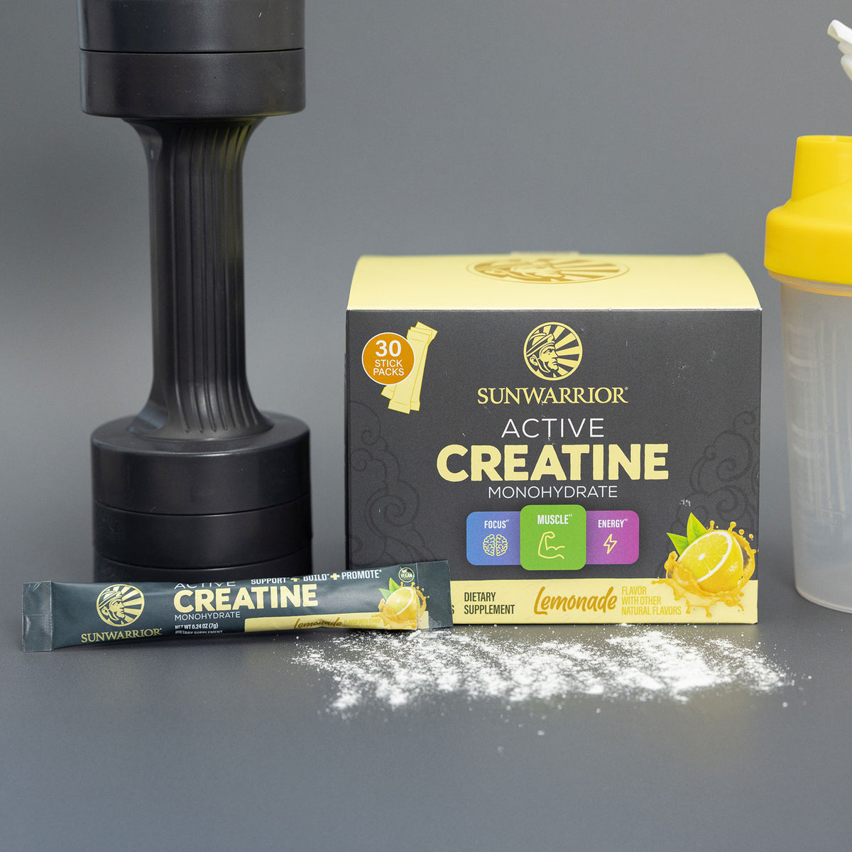 Sunwarrior Active Creatine Monohydrate lemonade flavor box with stick pack, shaker bottle, black dumbbell, and spilled powder on gray surface.