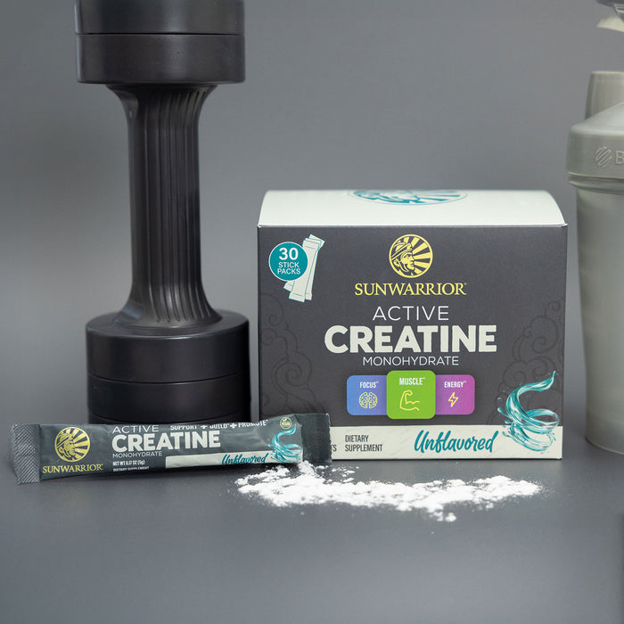 Sunwarrior Active Creatine monohydrate supplement box with an unflavored stick pack and spilled powder next to a black dumbbell and a white shaker bottle