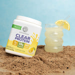 Container of lemon-flavored plant-based protein powder next to a glass of lemonade on sand.