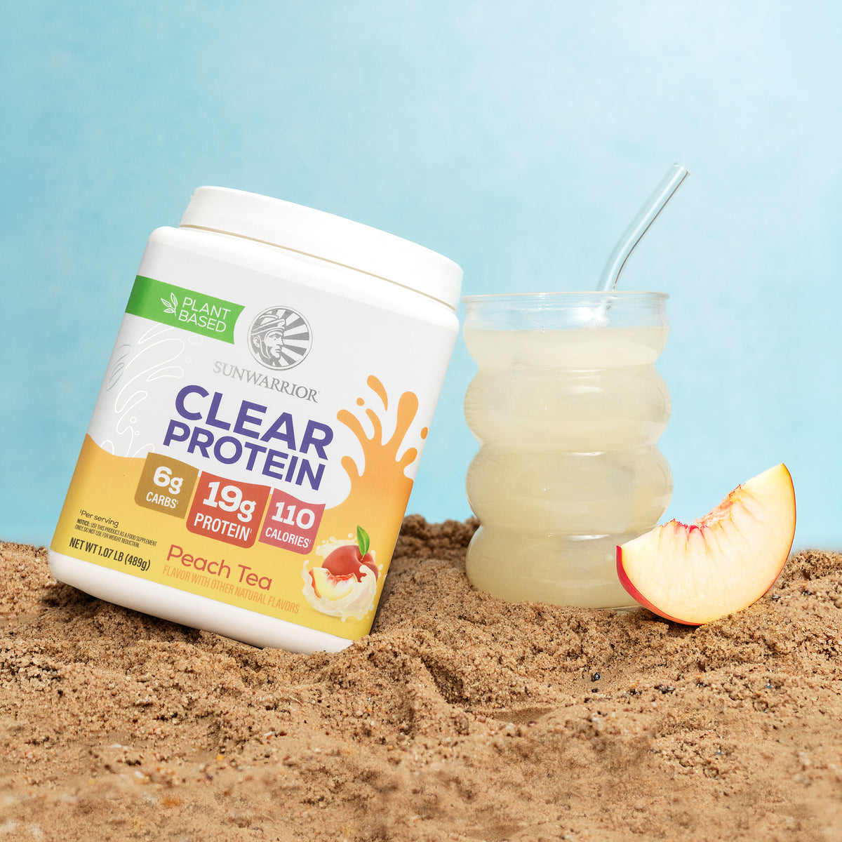 Sunwarrior Clear Protein Peach Tea container next to a glass of clear peach drink and a peach slice on sand