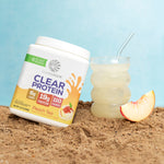 Sunwarrior Clear Protein Peach Tea container next to a glass of clear peach drink and a peach slice on sand