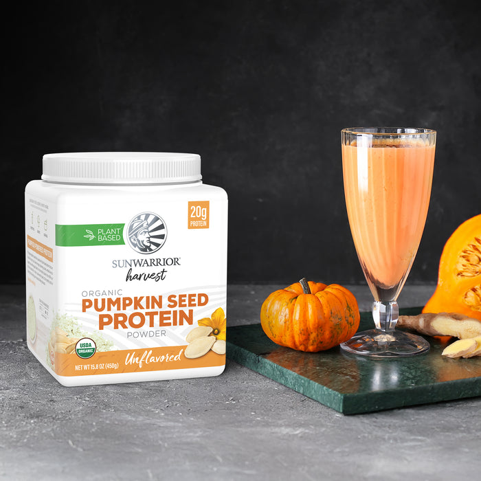 Container of organic pumpkin seed protein powder next to a small pumpkin and a filled smoothie glass