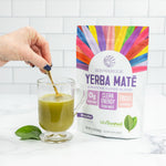 Hand stirring a green Yerba Mate and matcha tea in a glass cup beside a colorful Sunwarrior package