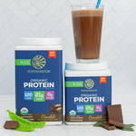 Two containers of plant-based chocolate protein powder with a glass of chocolate smoothie and pea pods on a white surface