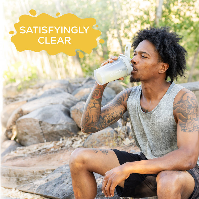 Man with tattoos wearing gray tank top drinking from a shaker bottle outdoors by rocks